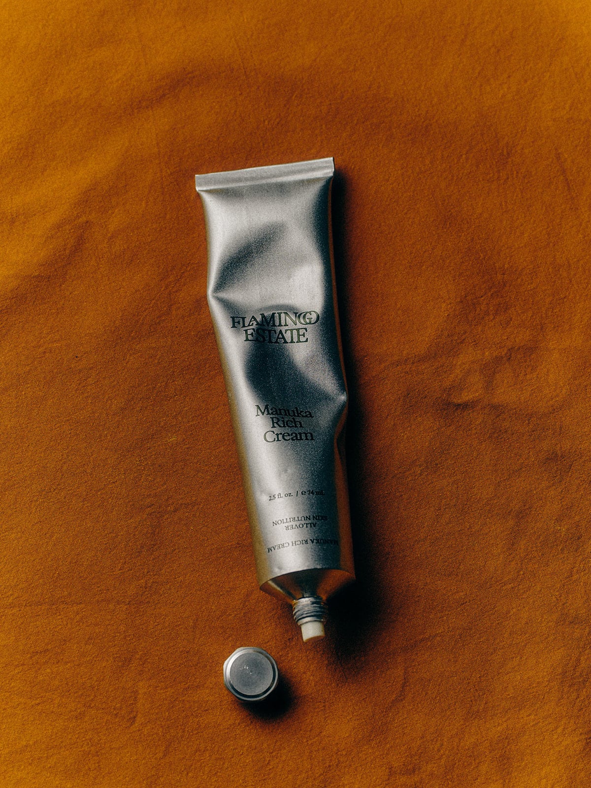 A silver tube of Flamingo Estate Manuka Rich Cream with the cap off, placed on a warm rust-colored fabric background.