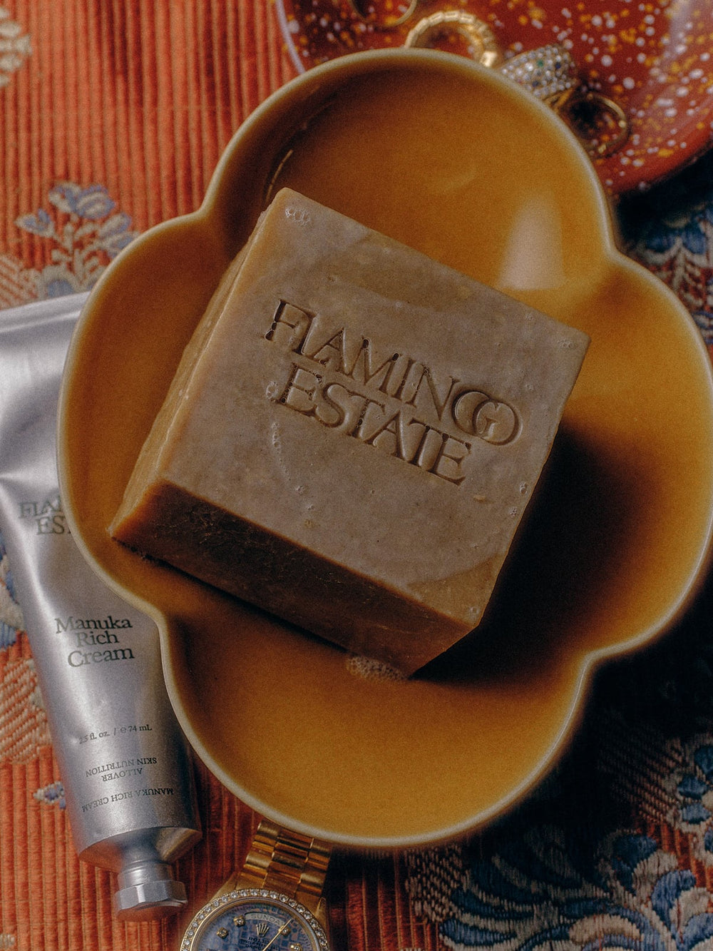A bar of Flamingo Estate Manuka Honey Soap in a mustard-yellow dish, with a tube of Manuka Rich Cream, a gold watch, and colorful fabric in the background.