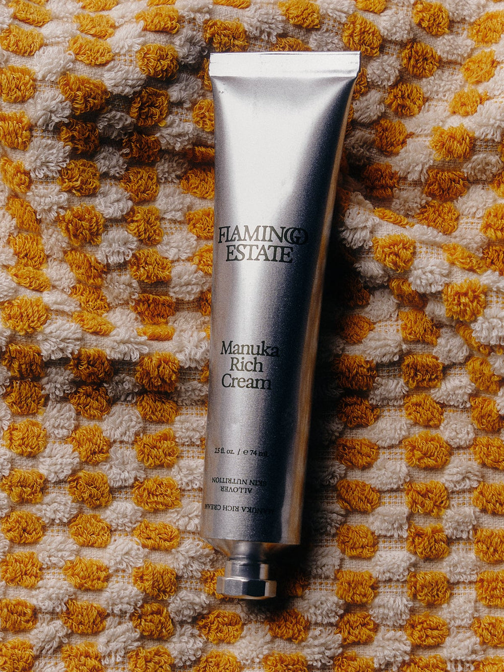 A silver tube of Flamingo Estate Manuka Rich Cream placed on a textured yellow and white woven towel.