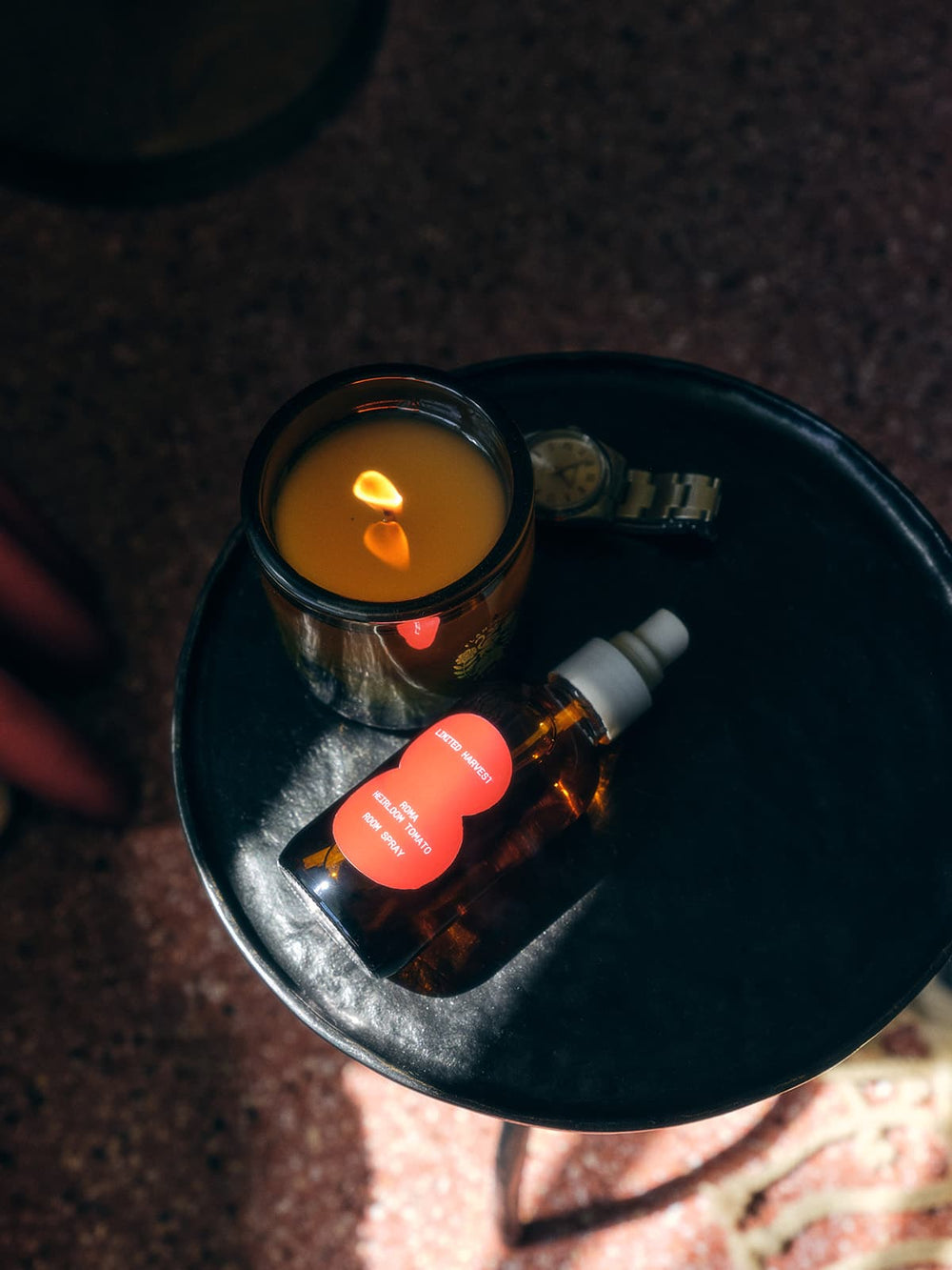 A lit Roma Heirloom Tomato candle and a Roma Heirloom room spray placed on a round black side table, creating a cozy and intimate ambiance.