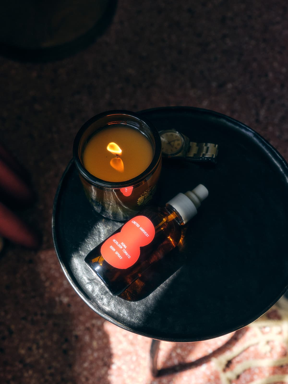A lit Roma Heirloom Tomato candle and a Roma Heirloom room spray placed on a round black side table, creating a cozy and intimate ambiance.