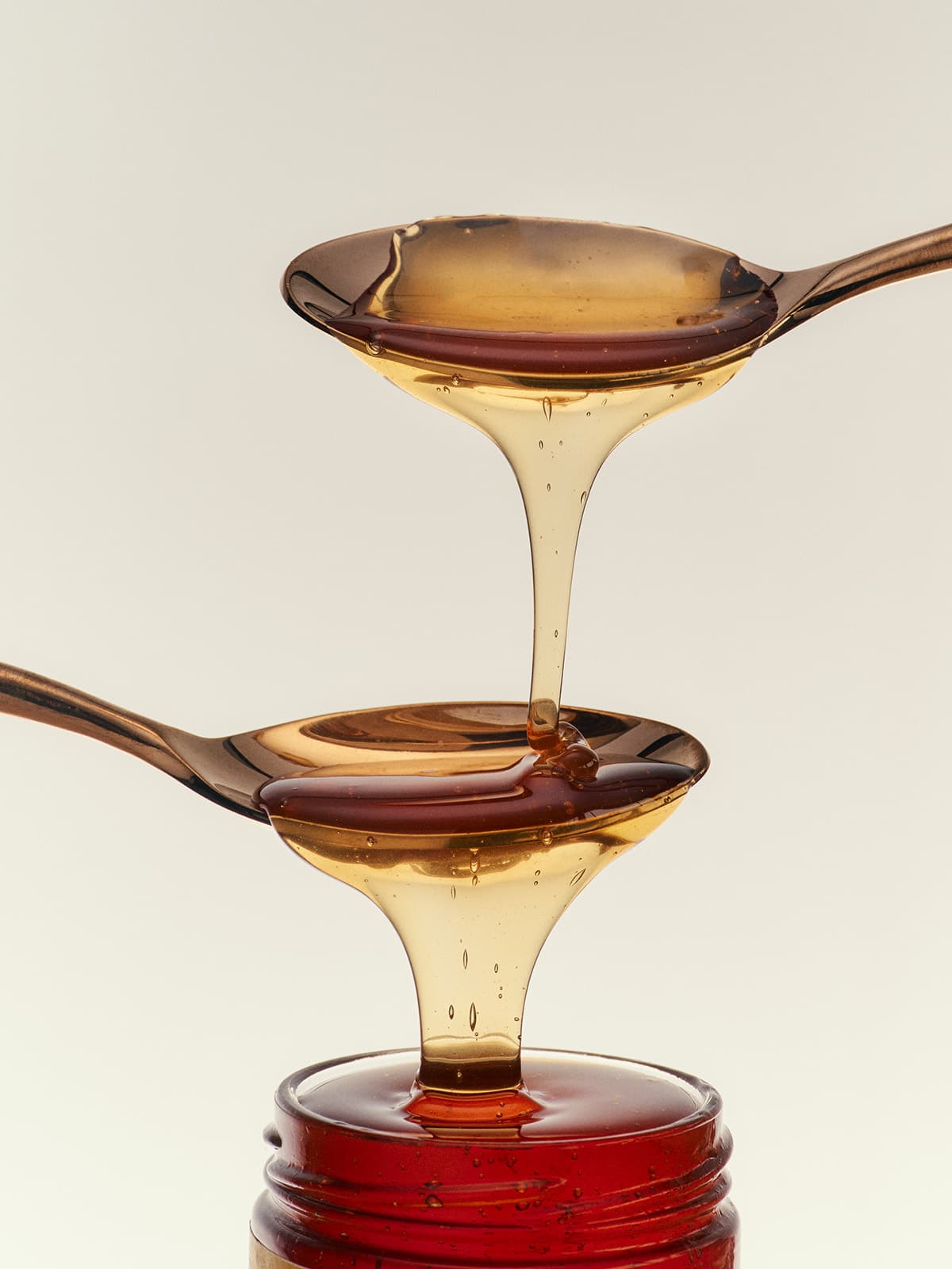 A spoon drips honey onto another lower spoon, positioned above a glass jar.