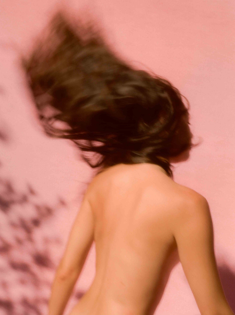 A blurred, nude figure with long, dark hair moves dynamically against a pink backdrop.