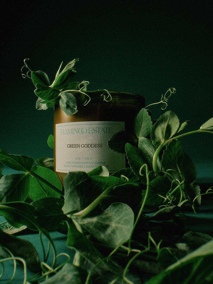 Candle labeled 'Green Goddess' from 'Farming Estate' surrounded by green leaves on a dark background