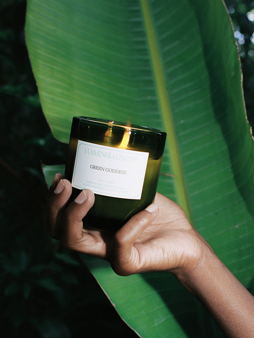 Hand holding a candle labeled 'Green Goddess' against a green leaf background