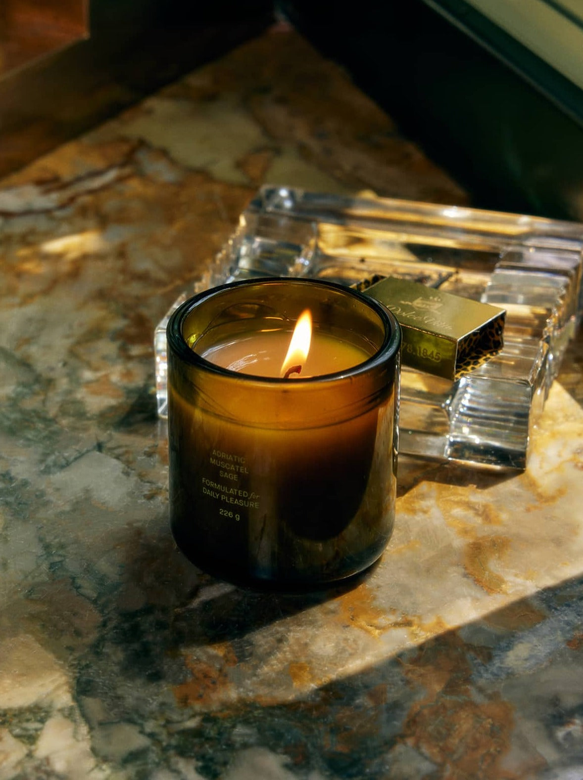 Lit Flamingo Estate Candle On A Marble Surface With A Glass Ashtray And A Box Of Matches