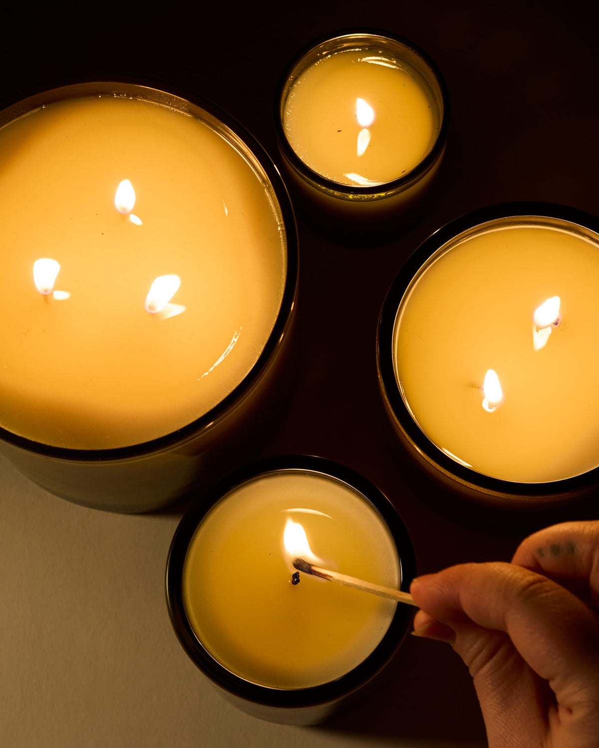 Close-up of Roma Heirloom tomato candles in black containers being lit by a match, highlighting the glow of the flame.