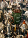 A green mug filled with coffee beans sits among various silver coffee pots on a reflective surface.