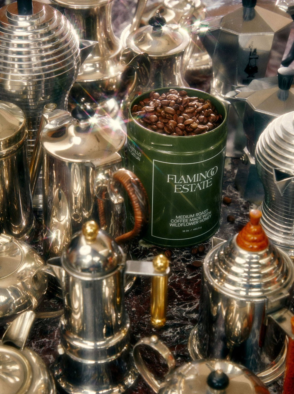 A green mug filled with coffee beans sits among various silver coffee pots on a reflective surface.