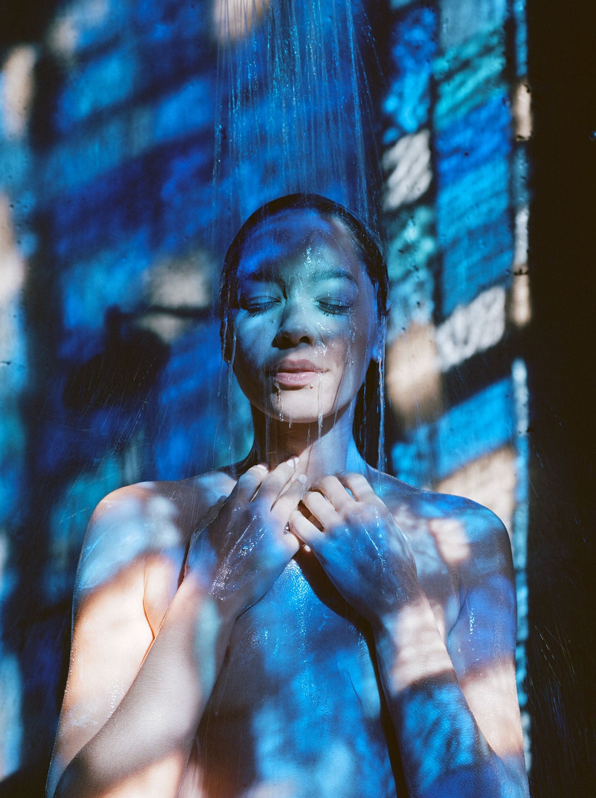 A person stands under a stream of water, eyes closed, hands near the chest.
