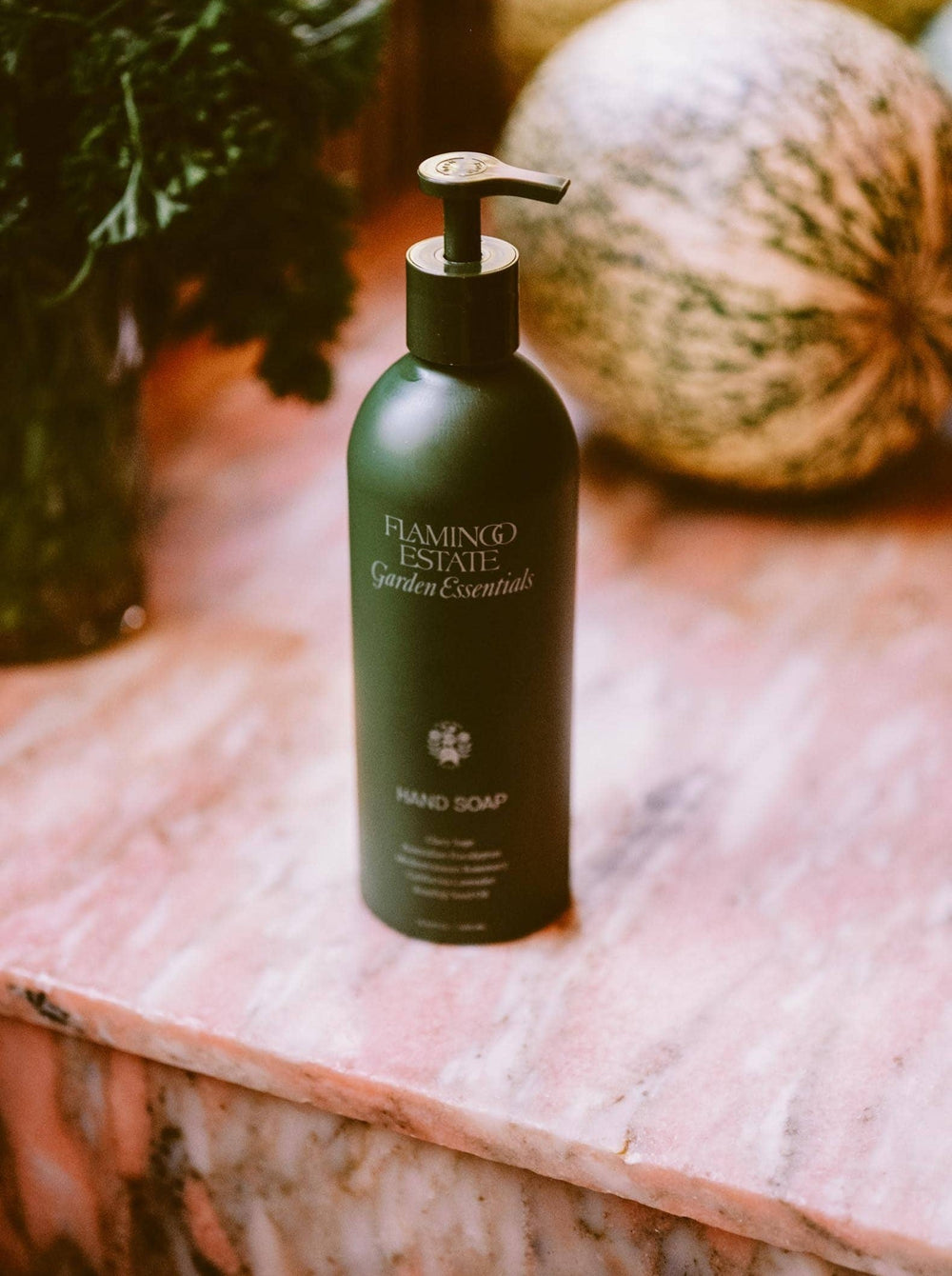 A Bottle Of Flamingo Estate Garden Essentials Handwash Behind A Sink, Next To Herbs And Watermelons